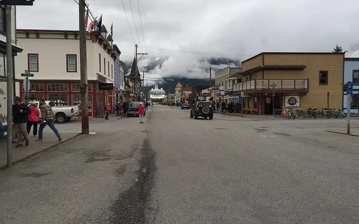 Broadway in Skagway lined with historic wooden storefronts, with a cruise ship and cloud-covered mountains at the end of the street