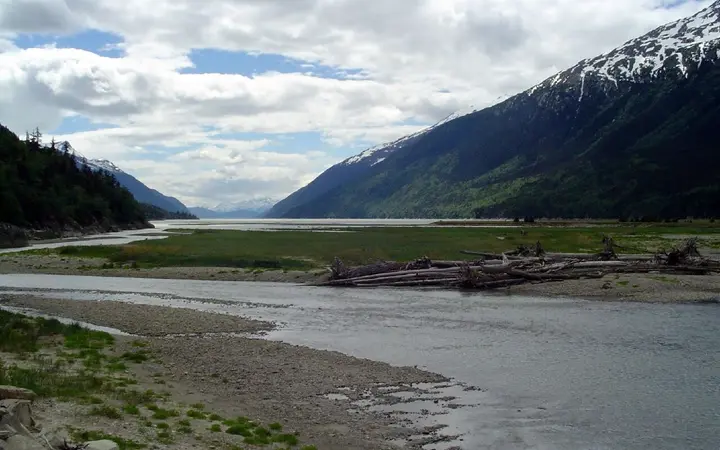 Tidal flats and river channel at the Dyea townsite with driftwood and snow-streaked mountains along Taiya Inlet