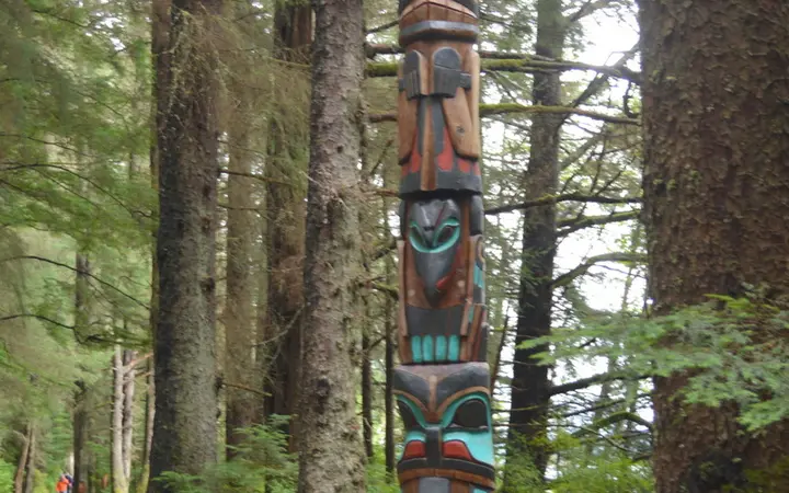 Carved totem pole beside a gravel path among tall spruce trees on the Totem Trail, Sitka National Historical Park