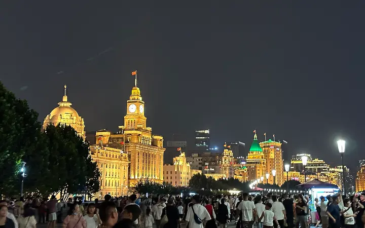 The Bund promenade at night with crowds in front of illuminated historic buildings including the Customs House clock tower
