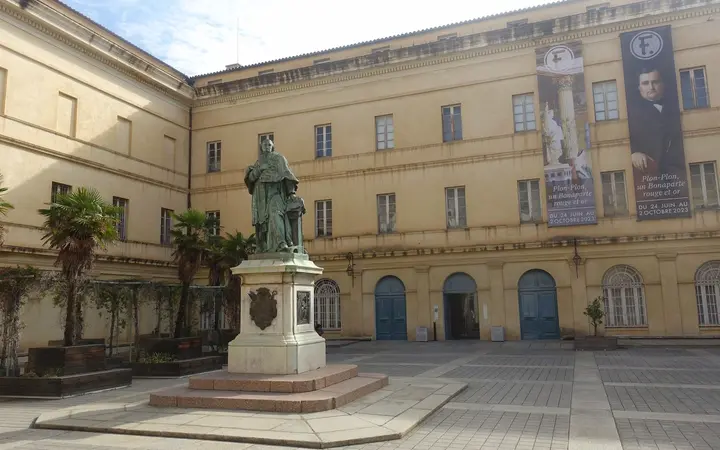Courtyard of the Palais Fesch in Ajaccio with the bronze statue of Cardinal Fesch on a pedestal in front of the ochre museum building, on a sunny day