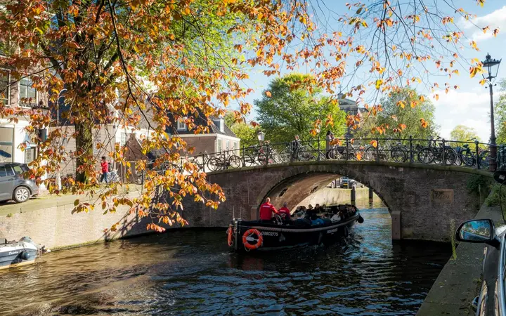 Arched stone bridge over the Reguliersgracht canal in Amsterdam on a sunny autumn day, with a canal boat passing under it and orange leaves in the foreground