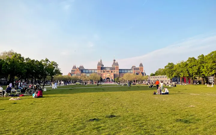The Rijksmuseum at the far end of the green Museumplein lawn under a clear blue sky, with trees on both sides and people relaxing on the grass in the distance