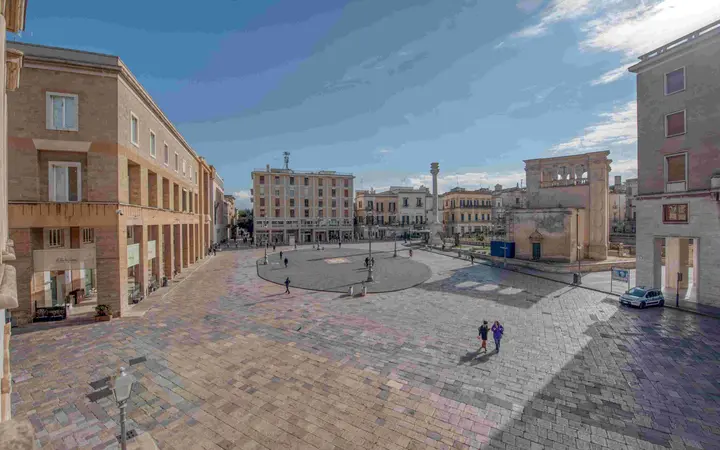 Wide view of Piazza Sant'Oronzo in Lecce with its paved square, old buildings and a few passers-by under a partly cloudy blue sky