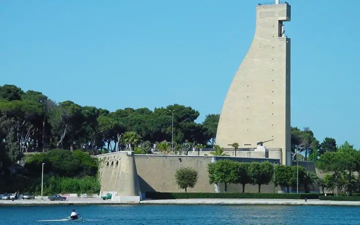 The tall rudder-shaped Monumento al Marinaio d'Italia above pine trees across Brindisi harbour, with a rower on the water under a clear blue sky