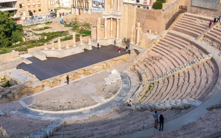 Daylight view over the restored seating and stage of the Roman Theatre of Cartagena, with old-town buildings behind