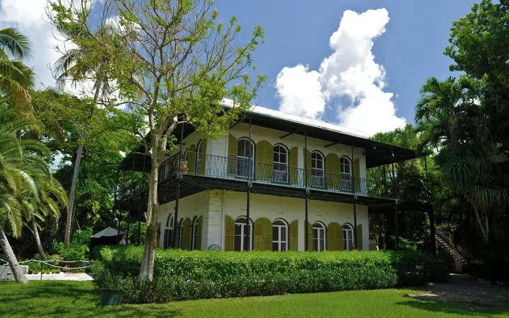 The white two-storey Hemingway House in Key West with green shutters, wraparound iron balconies and tropical gardens under a blue sky