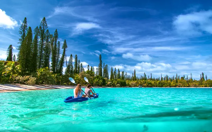 Kayakers on turquoise water in Kanuméra Bay beneath tall columnar pines.