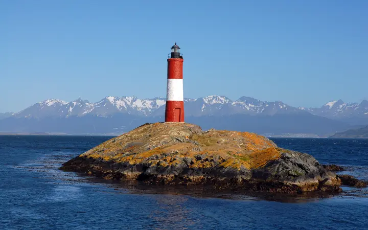 The red-and-white Les Eclaireurs lighthouse on a rocky islet in the Beagle Channel, with snowy peaks behind.