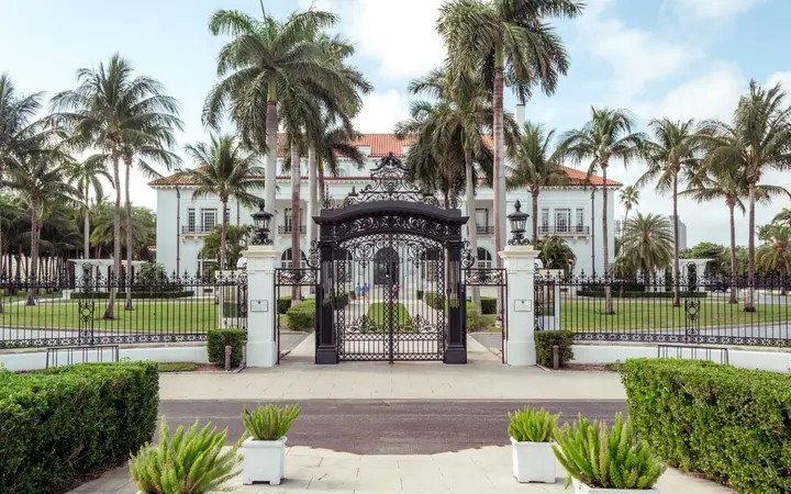 The ornate iron entrance gates and palm-lined drive of the Flagler Museum (Whitehall) in Palm Beach on a sunny day.