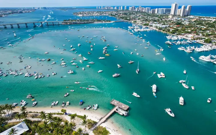 Aerial view of Peanut Island's beach and the turquoise Lake Worth Lagoon, with boats anchored offshore and the Singer Island skyline behind.
